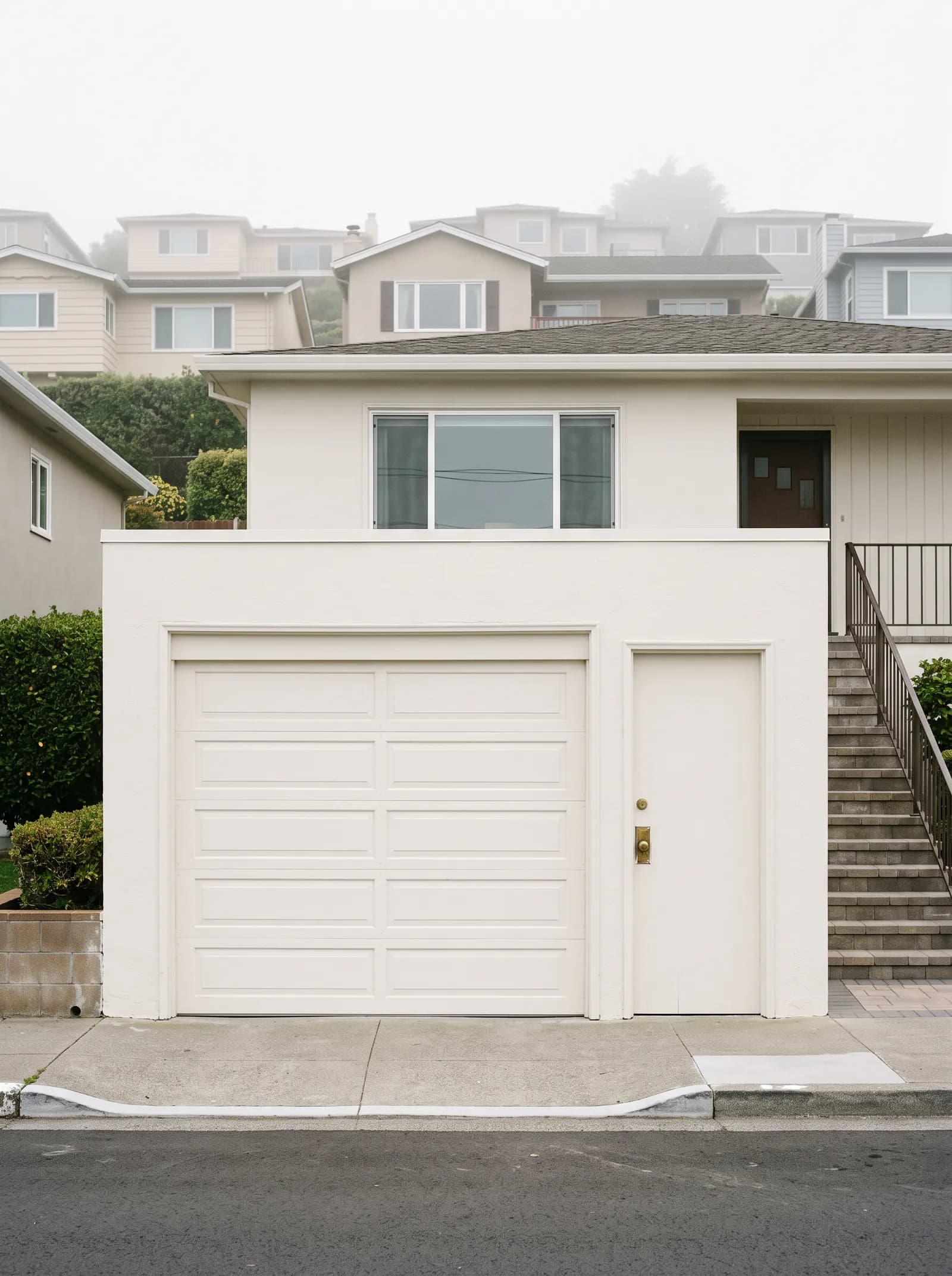 A Westlake house from the pavement: the single-car garage door at street level, the narrow door into the same garage beside it with a brass deadbolt at hand height, the living floor and its window above, and the steps climbing to the front door nobody uses.